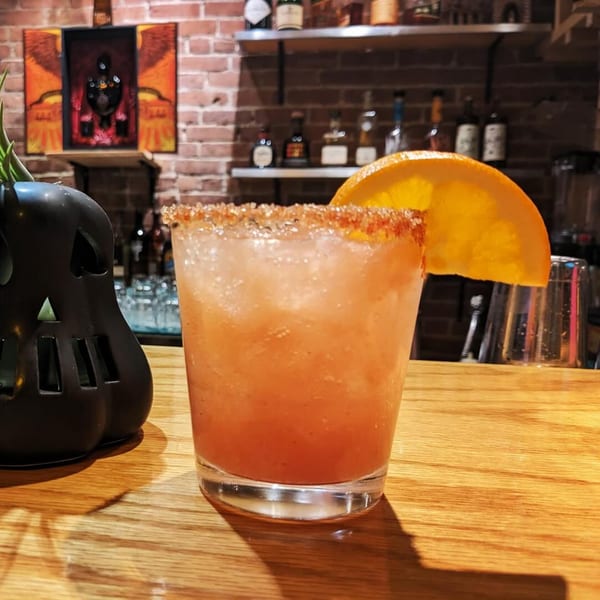 A Sol Azteca cocktail with a spiced rim and orange slice sits on a wooden bar next to a black skull decoration.