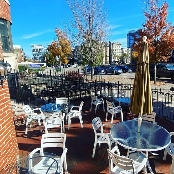 Outdoor patio with white chairs, glass tables, and a closed umbrella on a sunny day.