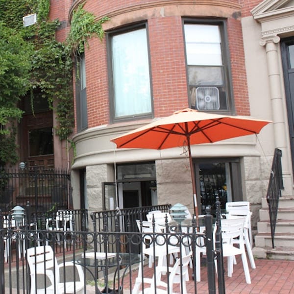 Sol Azteca's outdoor patio with white chairs, black tables, and an orange umbrella in front of a brick building.